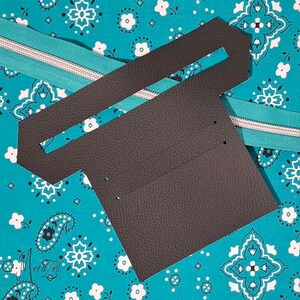 Puede incluir: Una pieza de cuero texturizado gris oscuro troquelada con una ranura horizontal y peque&ntilde;os orificios preperforados, colocada sobre una tela turquesa brillante con un patr&oacute;n de cachemira y flores blancas y una cremallera plateada.