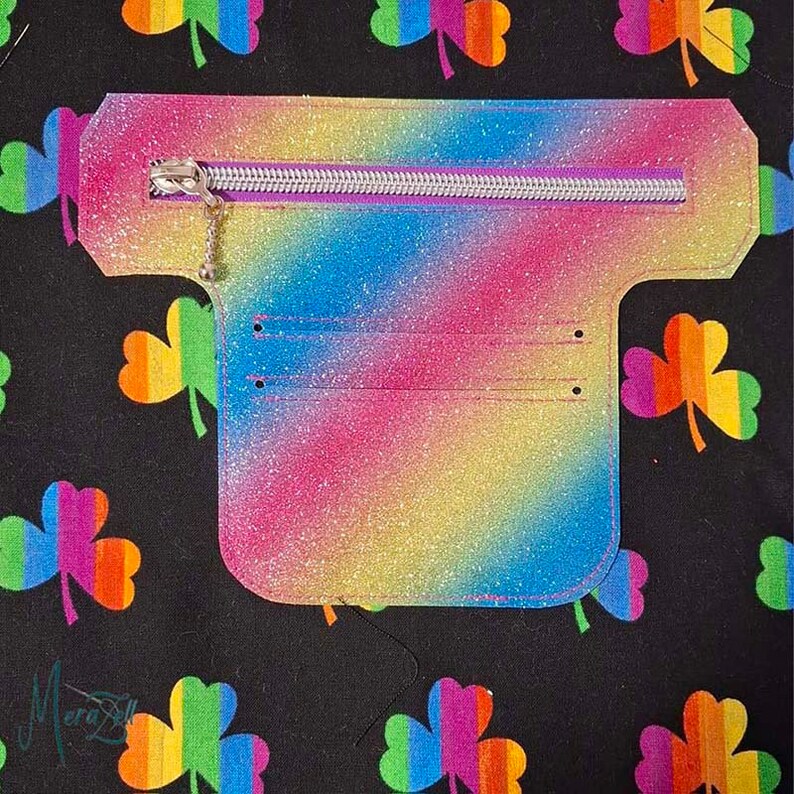 Puede incluir: Una cartera en forma de T con un acabado de purpurina arco&iacute;ris, que presenta una cremallera plateada en la parte superior y dos ranuras horizontales para tarjetas. La cartera se muestra sobre una tela negra con un patr&oacute;n de tr&eacute;boles arco&iacute;ris.