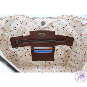Può includere: L'interno di una borsa tote con fodera beige e un motivo a piccole bacche rosse e blu. Include un pannello in pelle marrone con tasca con cerniera, due scomparti per carte contenenti due carte e una chiusura magnetica a scatto in alto.