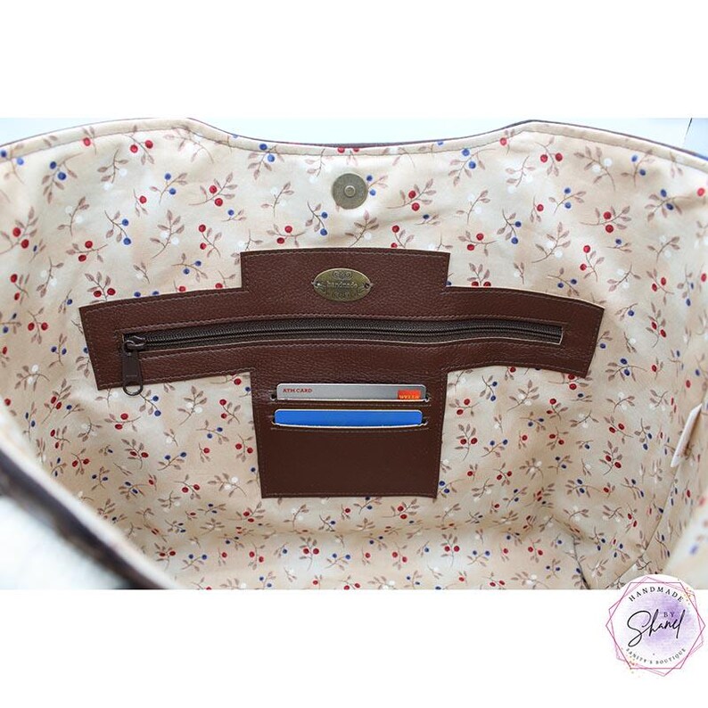 Puede incluir: El interior de un bolso tote con forro beige y un patr&oacute;n de peque&ntilde;as bayas rojas y azules. Incluye un panel de cuero marr&oacute;n con un bolsillo con cremallera, dos ranuras para tarjetas con dos tarjetas y un cierre magn&eacute;tico en la parte superior.