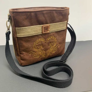 May include: A brown leather crossbody bag with a beige stripe and a gold embroidered heart design. The bag has a black adjustable strap and a small leather tag with the number "118" on it.