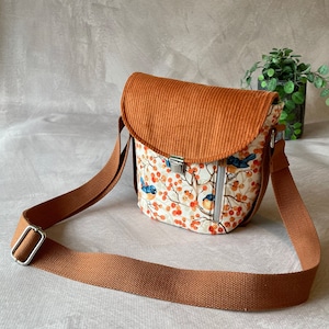 Op de afbeelding: Kleine crossbodytas met een oranje corduroy klep. De tas heeft een patroon met vogels, oranje bessen en takken op een witte achtergrond. Het heeft een bruine verstelbare riem en een rits.