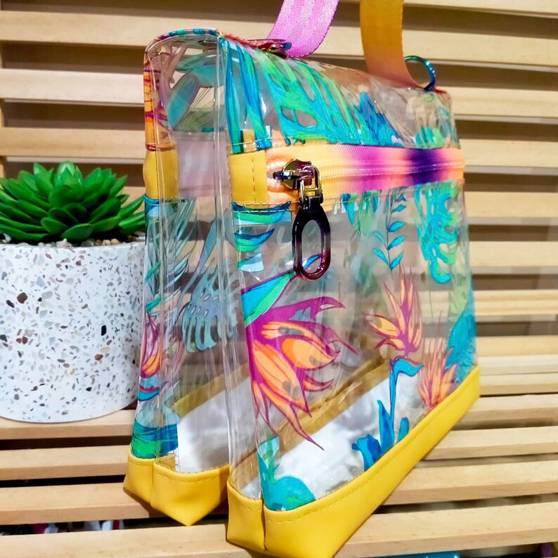 May include: A clear plastic bag with a yellow trim and a tropical print. The bag has a zipper closure and a pink and purple strap.