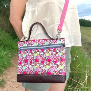 May include: A pink and white crossbody bag with a clear vinyl front featuring a repeating pattern of hearts, clouds, and gift boxes. The bag has a brown leather trim and a pink adjustable strap.