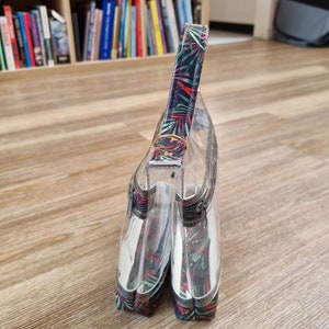 May include: Clear plastic double zippered pouch with a tropical leaf print handle. The pouch is standing upright on a wood floor.