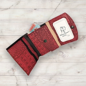 Puede incluir: Cartera de cuero roja con un patrón de cocodrilo en relieve. La cartera está abierta y tiene una ranura para tarjetas y un porta tarjetas de visita con el texto "Mera Bell" impreso en él.