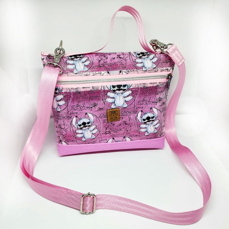 May include: Pink crossbody bag with a clear front panel featuring a repeating pattern of Stitch characters from the Disney movie Lilo & Stitch. The bag has a pink zipper and a pink adjustable strap.