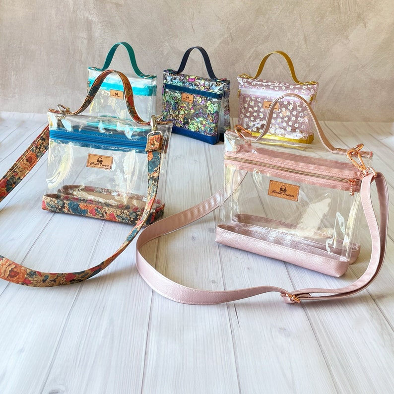May include: Five clear plastic crossbody bags with floral and patterned fabric lining. The bags have gold hardware and adjustable straps. The bags are arranged on a light brown surface.