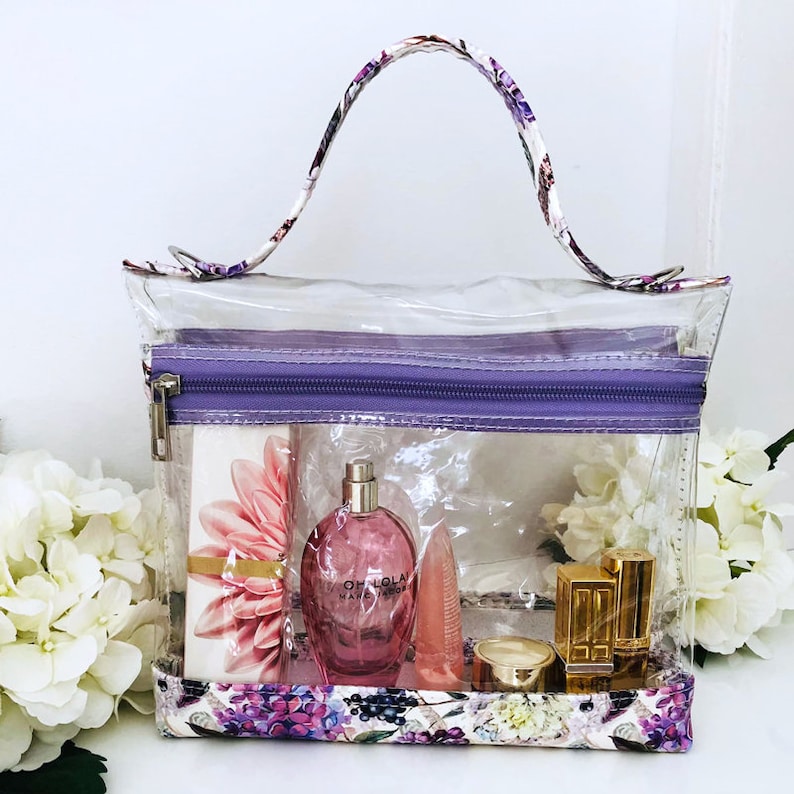 May include: Clear plastic zippered travel bag with a purple zipper and floral patterned trim. The bag contains a pink perfume bottle labeled "Oh Lola Marc Jacobs", a pink lip balm, a gold compact, and a gold lipstick.