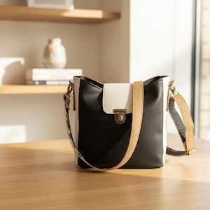 May include: A black, white, and tan leather shoulder bag with a flap closure and adjustable strap. The bag has a rectangular shape with a textured white flap and a tan strap. The bag is sitting on a wooden surface.