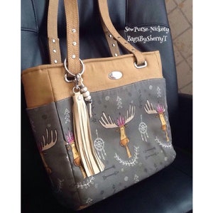 May include: A brown and grey tote bag with a moose print and a white tassel. The bag has a brown leather strap with silver studs and a silver ring. The text "Sew Purse-Nickety BagsBySherryT" is printed on the bag.