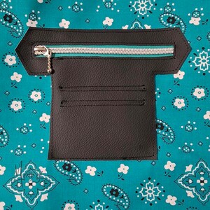 Puede incluir: Un panel de bolsillo de piel sint&eacute;tica negra con una cremallera horizontal plateada con ribete de tela verde azulado, unido a un fondo de tela verde azulado con un patr&oacute;n floral y de cachemira blanco. El bolsillo incluye dos ranuras.