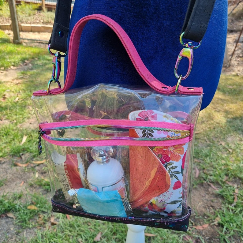 May include: A clear plastic tote bag with a pink handle and a black adjustable strap. The bag has a zippered compartment and is filled with various items, including a floral patterned mug, a bottle of perfume, and a blue washcloth.