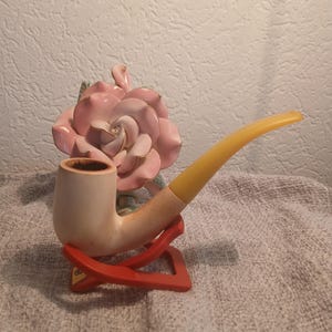 Puede incluir: Una pipa de tabaco con cazoleta de madera clara y boquilla amarilla, apoyada sobre un soporte plegable rojo. Detrás de la pipa hay una rosa decorativa de cerámica rosa con pétalos de borde dorado y hojas verdes en la base.