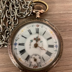 Op de afbeelding: Antieke zakhorloge met een zilveren ketting. De witte wijzerplaat heeft zwarte Romeinse cijfers en een kleine secondenwijzer. Delicate bloemmotieven in blauw, rood en goud sieren de wijzerplaat. Het horloge heeft een goudkleurige kroon en een zilveren kast.