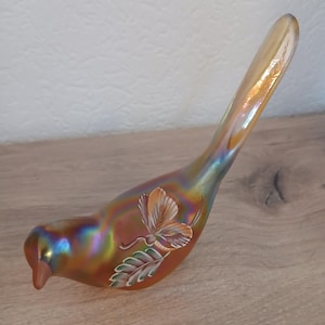 Könnte beinhalten: Eine irisierende Glasvogel-Figur mit langem Schwanz. Der Vogel ist hauptsächlich orange mit blauen und goldenen Akzenten. Ein gemalter Schmetterling und Blätter schmücken die Seite. Die Figur steht auf einer Holzoberfläche.
