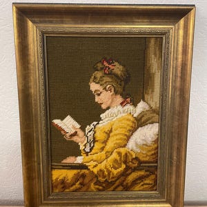 Könnte beinhalten: Gerahmtes Gobelin-Kunstwerk, das eine lesende Frau zeigt. Die Frau trägt ein gelbes Kleid mit weißem Kragen. Der Rahmen ist goldfarben, der Hintergrund dunkelgrün. Ein detailliertes, vintage Kunstwerk.