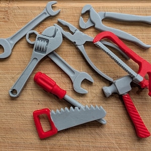 May include: A selection of toy tools, including spanners, pliers, a screwdriver, a hammer, and saws. The tools are mainly grey with red accents, arranged on a wooden surface.
