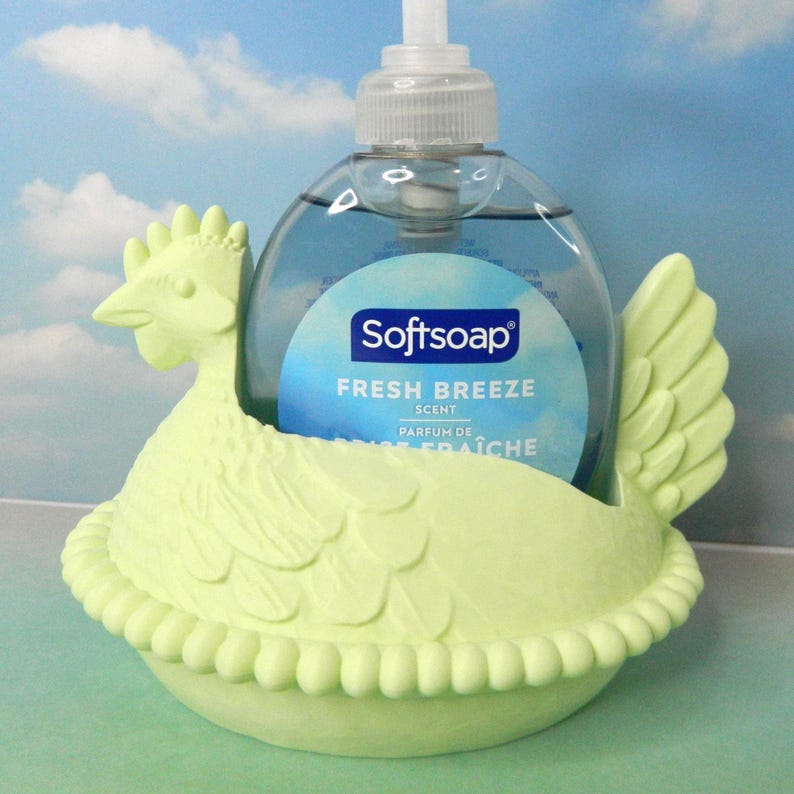 May include: A light green chicken-shaped soap dispenser holder. The holder is designed to cradle a clear bottle of Softsoap Fresh Breeze hand soap. The soap bottle has a blue label with white text. The background is a sky blue.