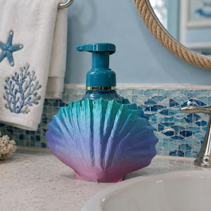 Pode incluir: Um dispensador de sabonete em forma de concha com um acabamento degradê que transita do azul-petróleo no topo para o roxo na base. O dispensador possui um mecanismo de bomba azul-petróleo no centro, projetado para conter sabonete líquido.