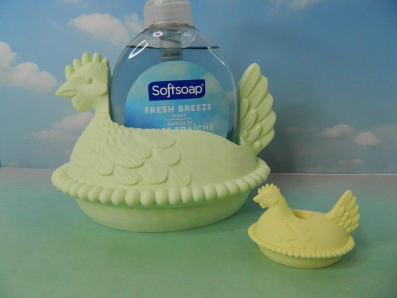 May include: A light green ceramic chicken-shaped soap dispenser holder with a matching small chicken-shaped soap dish. The holder contains a clear bottle of Softsoap Fresh Breeze scent. The background is a sky-blue color.