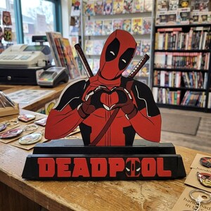 May include: A wooden cutout of the character Deadpool in a red and black suit, forming a heart shape with gloved hands. The figure stands on a black base with the red text DEADPOOL, featuring the character's mask icon as the letter O.