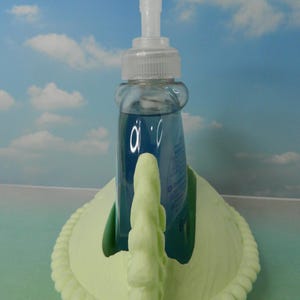 May include: A clear liquid soap dispenser with a white pump sits in a light green swan-shaped holder. The soap inside is blue. The holder has a decorative rope-like border.