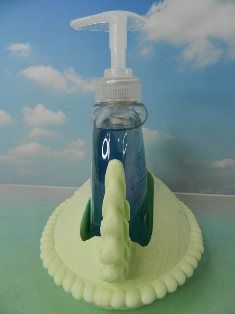 May include: A clear liquid soap dispenser with a white pump sits in a light green swan-shaped holder. The soap inside is blue. The holder has a decorative rope-like border.