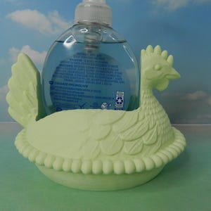 May include: A light green chicken-shaped soap dispenser holder. The holder is made of ceramic and has a detailed design. A clear plastic soap bottle with blue liquid is placed inside the holder.
