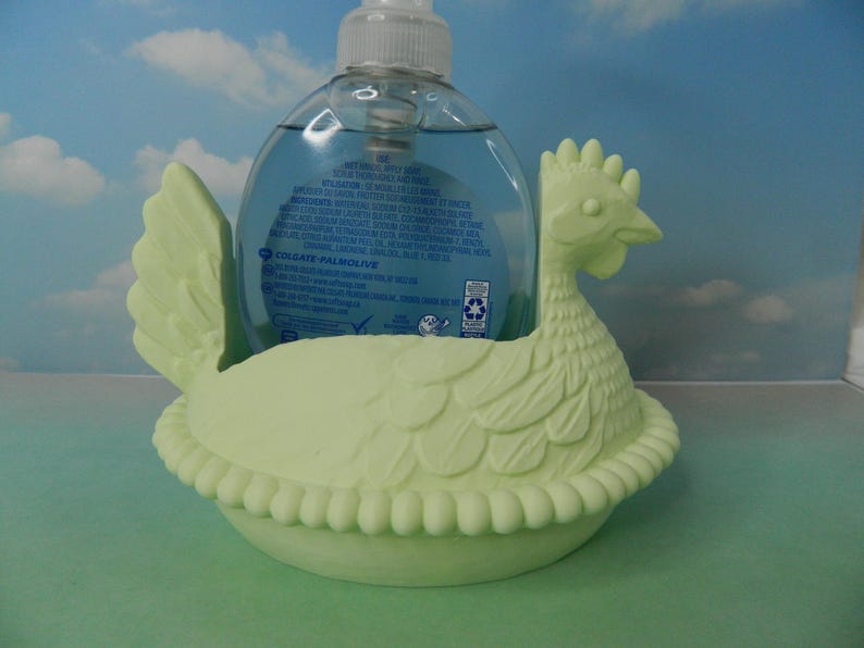 May include: A light green chicken-shaped soap dispenser holder. The holder is made of ceramic and has a detailed design. A clear plastic soap bottle with blue liquid is placed inside the holder.