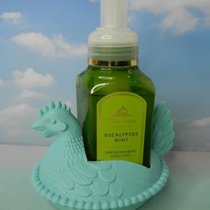Peut inclure: Un flacon de savon pour les mains vert avec une pompe blanche est plac&eacute; dans un support en forme de poulet bleu clair. L'&eacute;tiquette du savon indique "Eucalyptus Mint Gentle Foaming Hand Soap". Le support a un motif de plumes d&eacute;taill&eacute;.