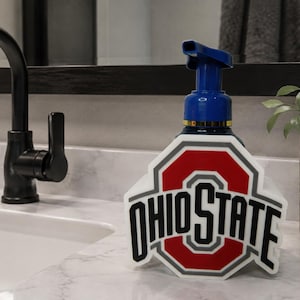 Könnte beinhalten: Ein blauer Seifenspender mit einem weißen, roten und schwarzen Ohio State-Logo auf der Vorderseite. Der Spender verfügt über einen Pumpmechanismus und ist für Flüssigseife vorgesehen, wobei das Branding deutlich auf der Oberfläche zu sehen ist.