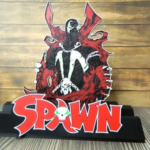 Puede incluir: Figura de Spawn, personaje de cómic en negro y rojo, sobre una base negra. La palabra "SPAWN" es roja con contorno blanco y un gráfico de calavera verde.