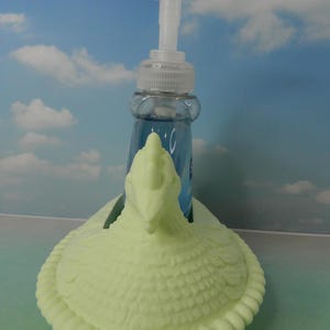 May include: A light green soap dispenser holder in the shape of a turkey. The holder has a textured body and a decorative rim. A clear plastic bottle with blue liquid and a white pump is inside the holder.