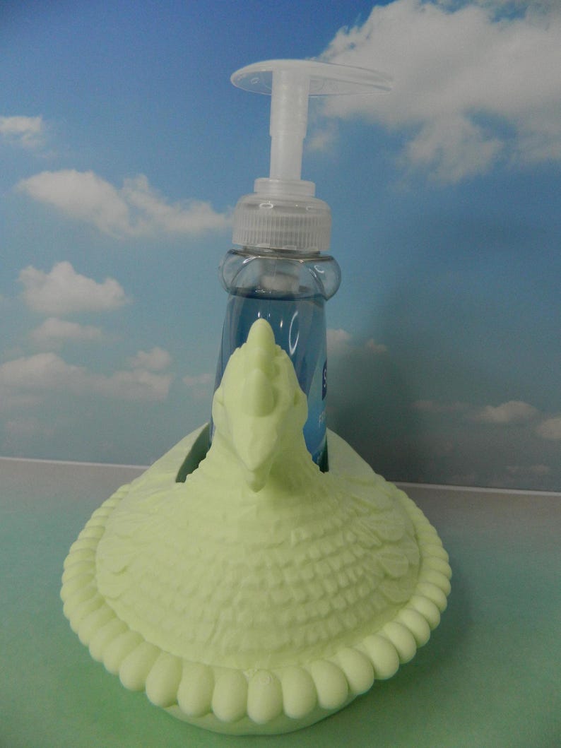 May include: A light green soap dispenser holder in the shape of a turkey. The holder has a textured body and a decorative rim. A clear plastic bottle with blue liquid and a white pump is inside the holder.