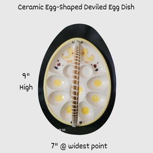 May include: Ceramic egg-shaped deviled egg dish, 9 inches high and 7 inches wide. The white dish has egg-shaped indentations with yellow centers, a woven handle, and a black border. The dish has a yellow rim and is decorated with duckling designs.