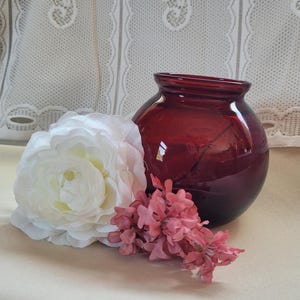 May include: A deep red glass vase with a round body and a wide opening. A large white flower and a small cluster of pink flowers are next to the vase. The background is a white lace curtain.