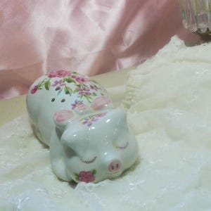 May include: A white ceramic pig figurine featuring a floral pattern of pink and purple flowers on its back and face. The pig has closed eyes with painted eyelashes, a pink snout, and small pink ears, depicted in a resting or sleeping position.