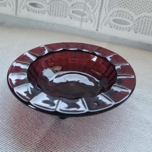 May include: A vintage, oval-shaped ashtray in a deep burgundy color. The ashtray has a glossy finish and a series of rectangular indentations around the rim. The interior is smooth and reflective, with a small, raised base.