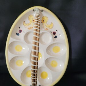 May include: Oval ceramic egg serving dish with a yellow rim and a woven handle. The dish has egg-shaped indentations with yellow yolks. Decorated with yellow ducklings and floral accents.
