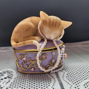 May include: A sleeping orange tabby cat figurine rests atop a purple jewelry box. The box is adorned with gold filigree, pink rhinestones, and a pearl necklace. The cat is curled up, wearing the necklace.