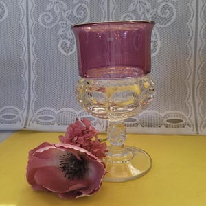May include: A clear glass goblet with a purple top and a decorative base. A pink flower rests near the goblet. The goblet has a gold rim and is likely used for serving beverages.
