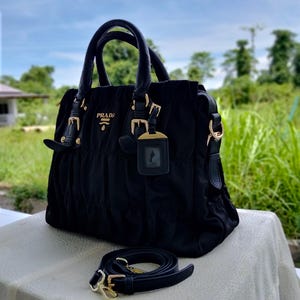 Puede incluir: Bolso de mano negro con herrajes dorados, con asa superior y correa de hombro ajustable. El bolso lleva el nombre de la marca "PRADA" en letras doradas. El fondo muestra un entorno natural al aire libre con follaje verde y cielo azul.