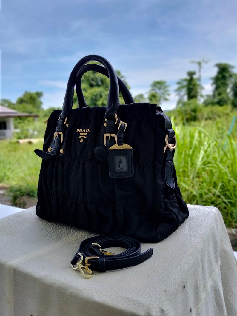 Puede incluir: Bolso de mano negro con herrajes dorados, con asa superior y correa de hombro ajustable. El bolso lleva el nombre de la marca "PRADA" en letras doradas. El fondo muestra un entorno natural al aire libre con follaje verde y cielo azul.