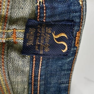 Puede incluir: Primer plano de tela vaquera con una etiqueta azul marino. La etiqueta presenta texto bordado en oro y un logotipo "S" estilizado. El denim tiene costuras naranjas y amarillas.