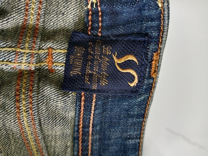 Puede incluir: Primer plano de tela vaquera con una etiqueta azul marino. La etiqueta presenta texto bordado en oro y un logotipo "S" estilizado. El denim tiene costuras naranjas y amarillas.