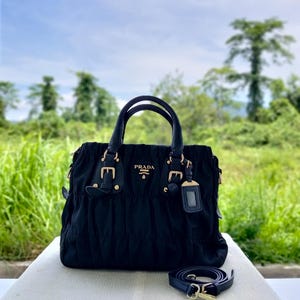 Puede incluir: Bolso Prada negro con herrajes dorados, con asa superior y bandolera extra&iacute;ble. El bolso tiene un dise&ntilde;o fruncido y un peque&ntilde;o logotipo de Prada en la parte delantera. El fondo muestra un campo verde y &aacute;rboles.