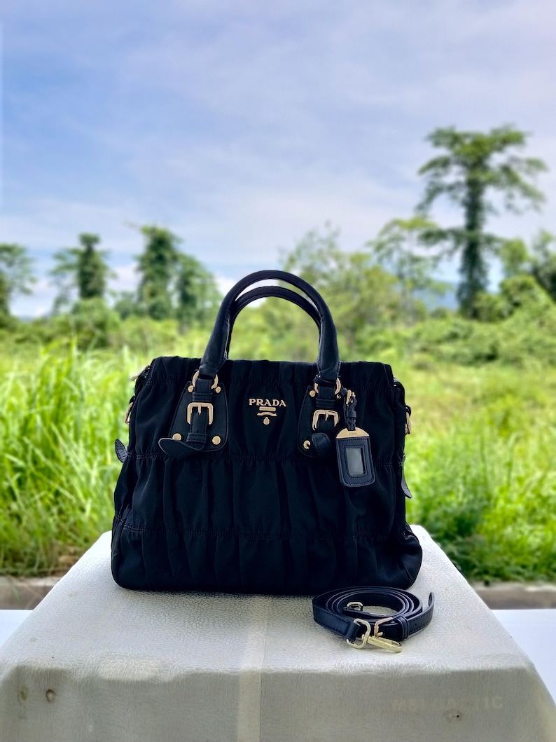 Puede incluir: Bolso Prada negro con herrajes dorados, con asa superior y bandolera extra&iacute;ble. El bolso tiene un dise&ntilde;o fruncido y un peque&ntilde;o logotipo de Prada en la parte delantera. El fondo muestra un campo verde y &aacute;rboles.
