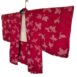 May include: A vibrant red kimono with a white patterned design. The traditional Japanese garment is hanging on a hanger, showcasing its long sleeves and intricate detailing. The kimono's fabric has a textured appearance.
