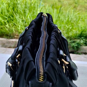 Puede incluir: Bolso de mano negro con cremallera y herrajes dorados. El bolso est&aacute; hecho de un material suave y fruncido, con detalles de cuero. La cremallera est&aacute; abierta, revelando el interior. El fondo es follaje verde borroso.